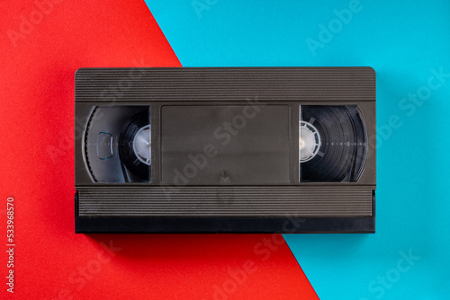 Black vintage VHS videotape cassette on red and blue background. Plastic retro video cassette with analog magnetic tape. Old technology for tape recording and watching media movies. Top view.
