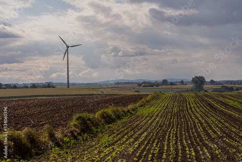 Wczesna jesień . Pola ze ściętą kukurydzą i i wysianym poplonem .Duży wiatrak ( turbina wiatrowa ) wytwarzający prąd . To wszystko na tle Gór Świetokrzyskich i nieba z chmurami .