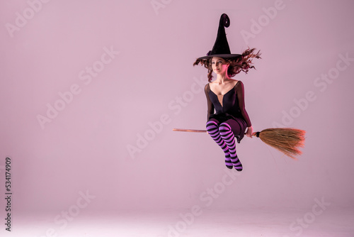 A ballerina on pointe shoes in a black witch costume in a hat flies on a broomstick on a lilac background. Ballet for Halloween. Copyspace.