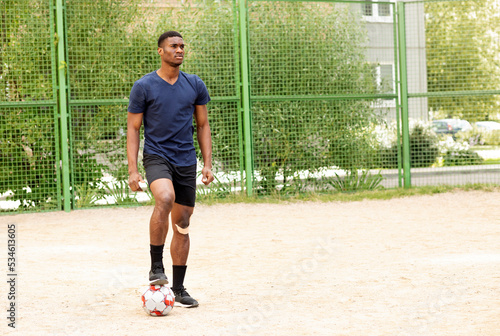 Young confident african american guy with taped knee holds soccer ball by foot outside, on stadium. Football lifestyle. Man plays the match,game. Big dream,future professional player. Horizontal plane