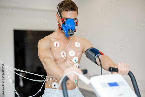 Man athlete with breath mask and electrodes training on bike simulator, examining his cardiovascular system at medical office