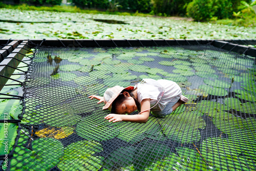 Asian little girl close her eyes and laying down on netting park inside lotus pond garden,relax time.Child on top net trampoline. Outdoor activities for kids.
