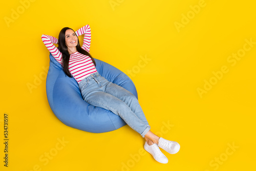 Full size photo of good mood girl wear striped long sleeve jeans lean back on pouf look empty space isolated on yellow color background