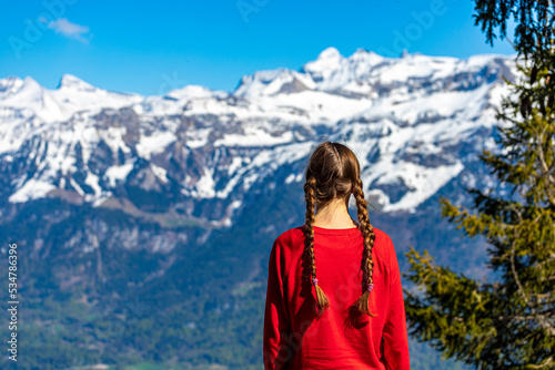 sweet woman with pigtails stands on top of a mountain in the swiss alps admiring the view of the snow-capped peaks; spring hike in the swiss alps, interlaken