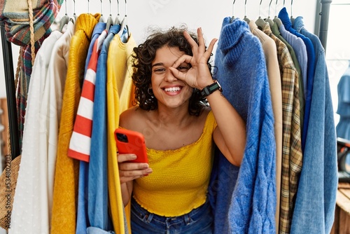 Young hispanic woman searching clothes on clothing rack using smartphone doing ok gesture with hand smiling, eye looking through fingers with happy face.