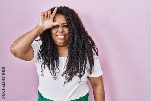 Plus size hispanic woman standing over pink background making fun of people with fingers on forehead doing loser gesture mocking and insulting.