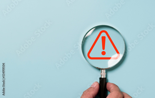 Hand holding magnifier glass with red triangle caution warning sign for focus notification error and maintenance concept.