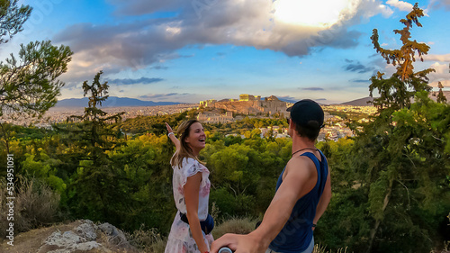 A young couple taking a selfie during sunset with panoramic view looking at Parthenon of the Acropolis of Athens during sunset seen from Filopappou Hill, Attica, Greece, Europe. Ruins ancient temple