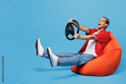 Full body young man of African American ethnicity 20s wear red shirt sit in bag chair hold steering wheel pretend driving isolated on plain pastel light blue cyan background. People lifestyle concept.