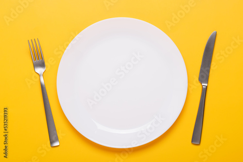 Clean empty white plate with knife and fork on yellow background place setting top view