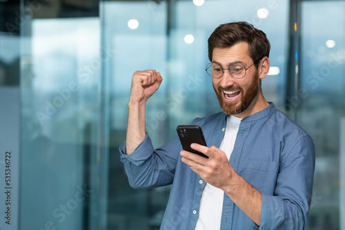 Successful businessman working in modern office building, man looking at smartphone and reading news celebrating happy news victory businessman in shirt and glasses holding hand up triumph gesture.