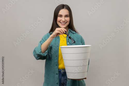 Smiling woman with dark hair standing holding trash bin and throwing out eyeglasses, improving eyesight after treatment, wearing casual style jacket. Indoor studio shot isolated on gray background.