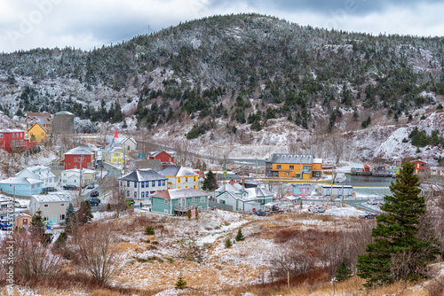 A small fishing village, Quidi Vidi Village, in Newfoundland. Colorful wooden houses surround a small fishing harbor. The aerial view shows snow on the ground and on the tree covered hill. 