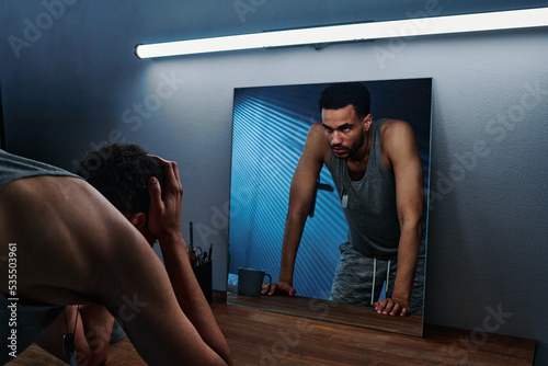 Young insomniac man holding head in hands in front of mirror with reflection of him expressing anger and aggression