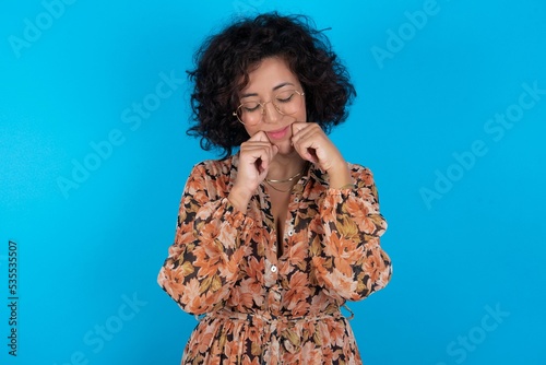 Pleased young brunette woman with curly hair wearing flowered dress standing over blue background with closed eyes keeps hands near cheeks and smiles tenderly imagines something very pleasant