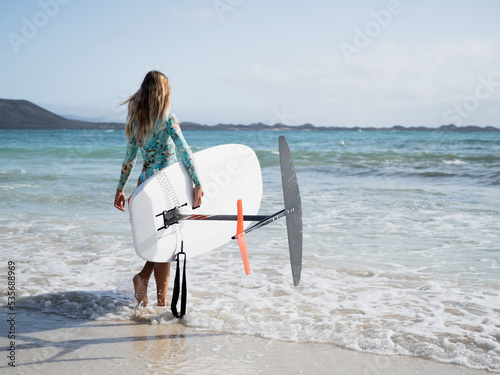 Young woman from behind walking to the water on the beach shore to practice hydrofoil surfing water sport