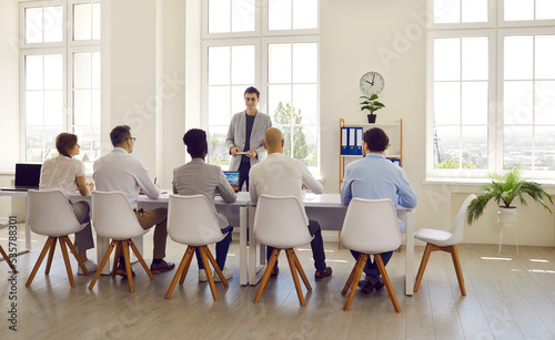 Man talks about himself while speaking in front of human resources interview committee in office. Rear view of people sitting in row and listening to young man's speech. Employment and hiring concept.