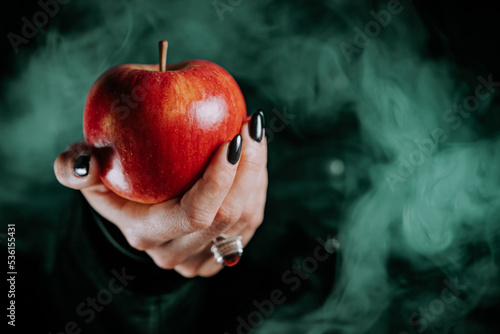 Woman as witch offers red apple as symbol of temptation, poison. Fairy tale, white snow wizard concept. Spooky halloween, cosplay. Smoke, haze background.
