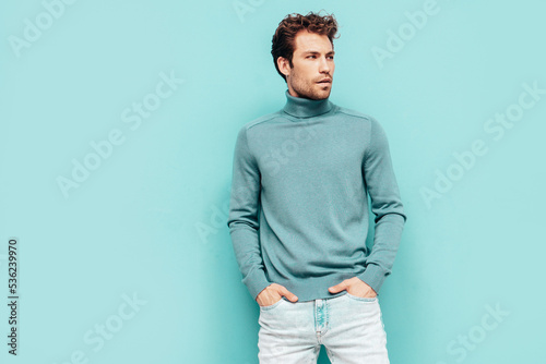 Portrait of handsome confident model. Sexy stylish man dressed in sweater and jeans. Fashion hipster male with curly hairstyle posing near blue wall in studio. Isolated