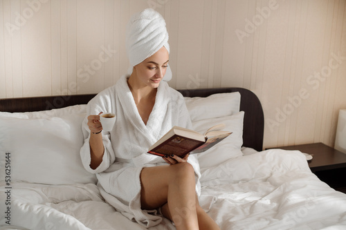 Pretty caucasian female in bathrobe and towel on head is reading a book on the bed