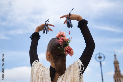 Portrait of young teenage girl in black dance dress, white shawl and pink carnations in her hair, dancing flamenco with castanets in her hands. Concept of flamenco, dance, art, typical Spanish dance.