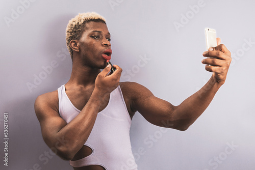 An androgynous black man posing in a purple studio while putting on lipstick while using his cell phone, LGTBI concept