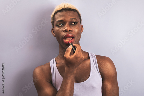 An androgynous black man posing against a purple studio background while putting on lipstick.
