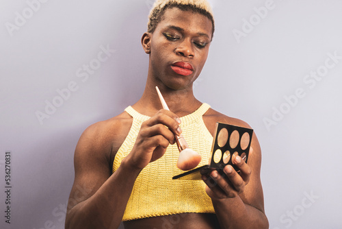An androgynous black guy posing on a purple studio background while using cosmetics.