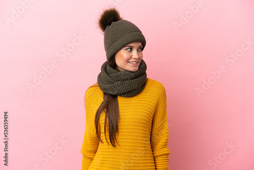 Young caucasian girl with winter hat isolated on pink background looking to the side and smiling