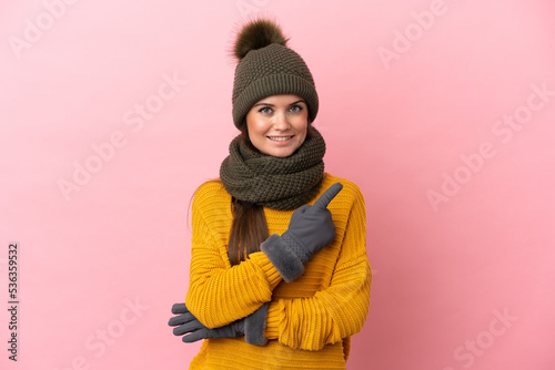 Young caucasian girl with winter hat isolated on pink background pointing to the side to present a product
