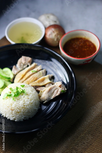 Hainanese chicken rice served on a black plate with dipping sauce and hot broth on white table.