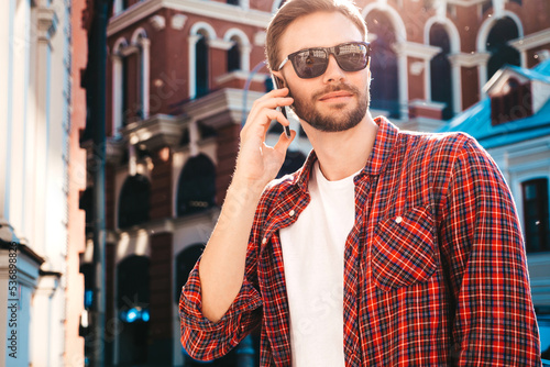 Handsome stylish hipster lambersexual model. Modern man dressed in red shirt. Fashion male posing on the street background in sunglasses. Outdoors at sunset. Using smartphone. Talking