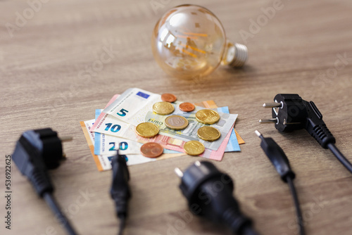 expensive electricity concept - electric power plugs, bulb and european money banknotes and coins on the table