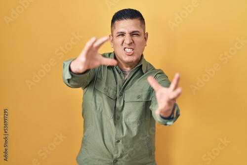 Hispanic young man standing over yellow background shouting frustrated with rage, hands trying to strangle, yelling mad