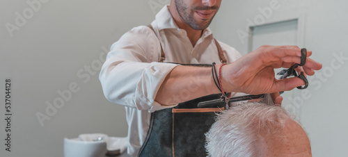 Hombre peluquero irreconocible, cortando el pelo con tijera, a cliente senior anónimo, en la peluquería. Fotografía horizontal con espacio para texto.