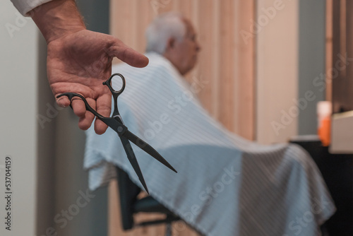 Detalle de mano de peluquero con tijeras, y de segundo plano, cliente senior, sentado para cortar el pelo.