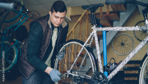 Experienced mechanic in gloves is assembling bicycle adjusting back wheel during servicing vehicle in workplace. Professional instruments, spare parts and equipment are visible.