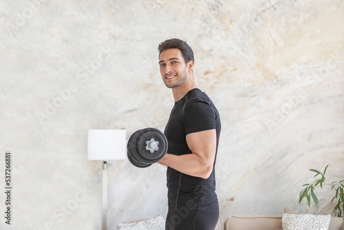 Side view of young handsome man in black sportswear doing biceps curl with dumbbells in living room at home. Healthy lifestyle and home workout concept