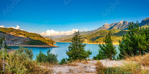 Beautiful picturesque panorama of a mountainous region with hills covered with forests, a lake and rugged mountains against a blue sky.
