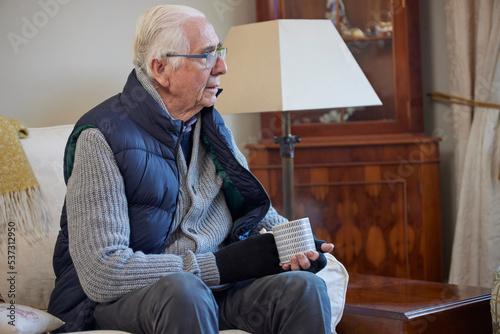 Senior Man Wearing Extra Clothes With Hot Drink Trying To Keep Warm At Home In Energy Crisis