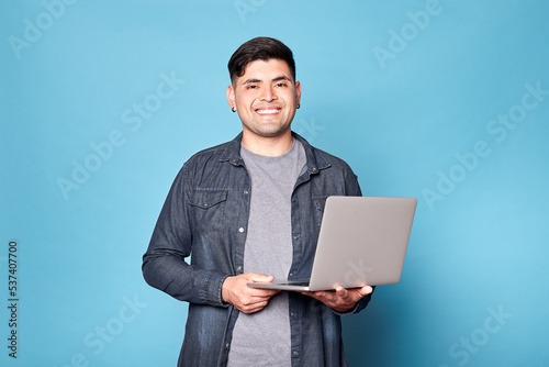 Portrait of a happy latino young man standing, holding laptop, isolated on blue background. Copy space.