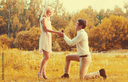 Happy young couple, man kneeling down and proposing a ring to his beloved woman outdoors in the park, wedding concept