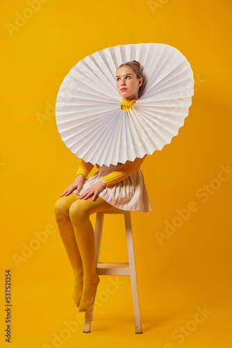 Young indifferent girl in giant jabot collar or neckwear and yellow tights isolated over yellow background. Contemporary art, weird beauty, fashion