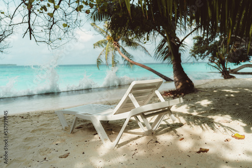 View of a daybed in front of the ocean on a luxury Maldives resort on a warm sunny day surrounded by palm trees; a plastic recliner on the coral sand in front of breaking waves on the beach