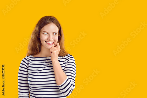 Young woman looks aside with funny awkward smile on face. Beautiful lady standing on yellow studio background looks at blank copy space side, sees something, thinks of guilty pleasure, feels nervous