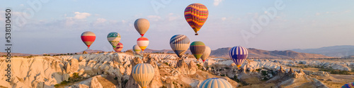 Colorful hot air balloons under rocky landscape of natural formations at sunrise in Cappadocia, central Turkey. Web banner header.