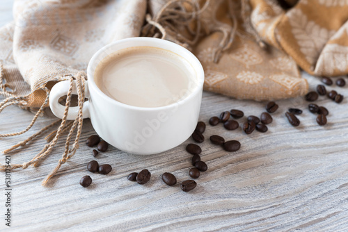 Cozy background with coffee cup, some scattered coffee beans and fabric scarf on the light wooden background. Copy space