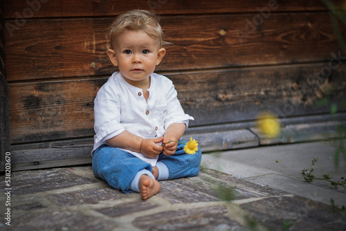one year old happy blond baby boy in white shirt sitting outside on the ground infront of a rustic wooden hut and holding a yellow marguerite flower to send love and happiness
