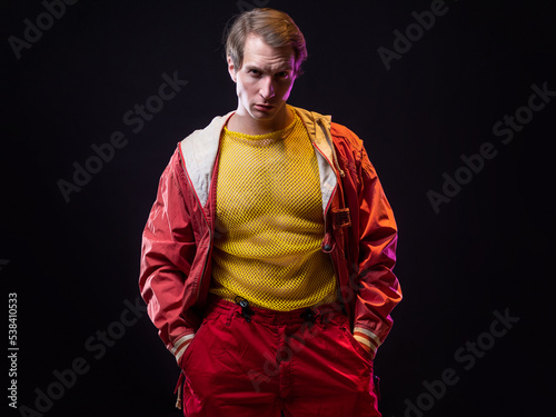 a young muscular man in a bright outfit poses in a pretentious pose, style and grotesque. Attractive guy with pumped-up arms in a mesh T-shirt and red pants, photo in smoke