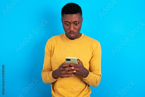 Focused young handsome man wearing yellow T-shirt over blue background use smartphone reading social media news, or important e-mail
